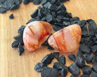 Puede incluir: Un par de pendientes de botón con una forma semicircular, lisa y redondeada. Los pendientes presentan una base de color rosa pálido translúcido con vetas de color rojo oscuro y escamas metálicas doradas incrustadas en la superficie.