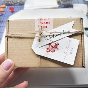 Puede incluir: Una caja rectangular de cart&oacute;n marr&oacute;n atada con cordel de yute fino. Incluye una pegatina rosa con el texto THE PACKAGE IS HAPPY y dos etiquetas de papel blanco. Una etiqueta dice Valentines Day y la otra muestra corazones rojos y el texto DAY.