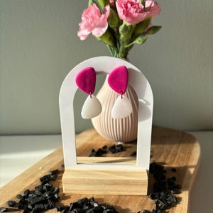 Könnte beinhalten: Ein Paar Hängeohrringe mit einem leuchtend fuchsiafarbenen, halbkreisförmigen Oberteil, das mit einem weißen, durchscheinenden, abgerundeten dreieckigen Unterteil verbunden ist, präsentiert auf einem weißen bogenförmigen Ständer mit Holzsockel.