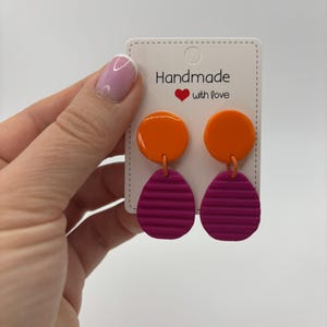 May include: Dangle earrings featuring a smooth, glossy orange circular top piece connected to a textured, matte magenta oval bottom piece with horizontal ridges. The earrings are attached to a white display card labeled Handmade with love.