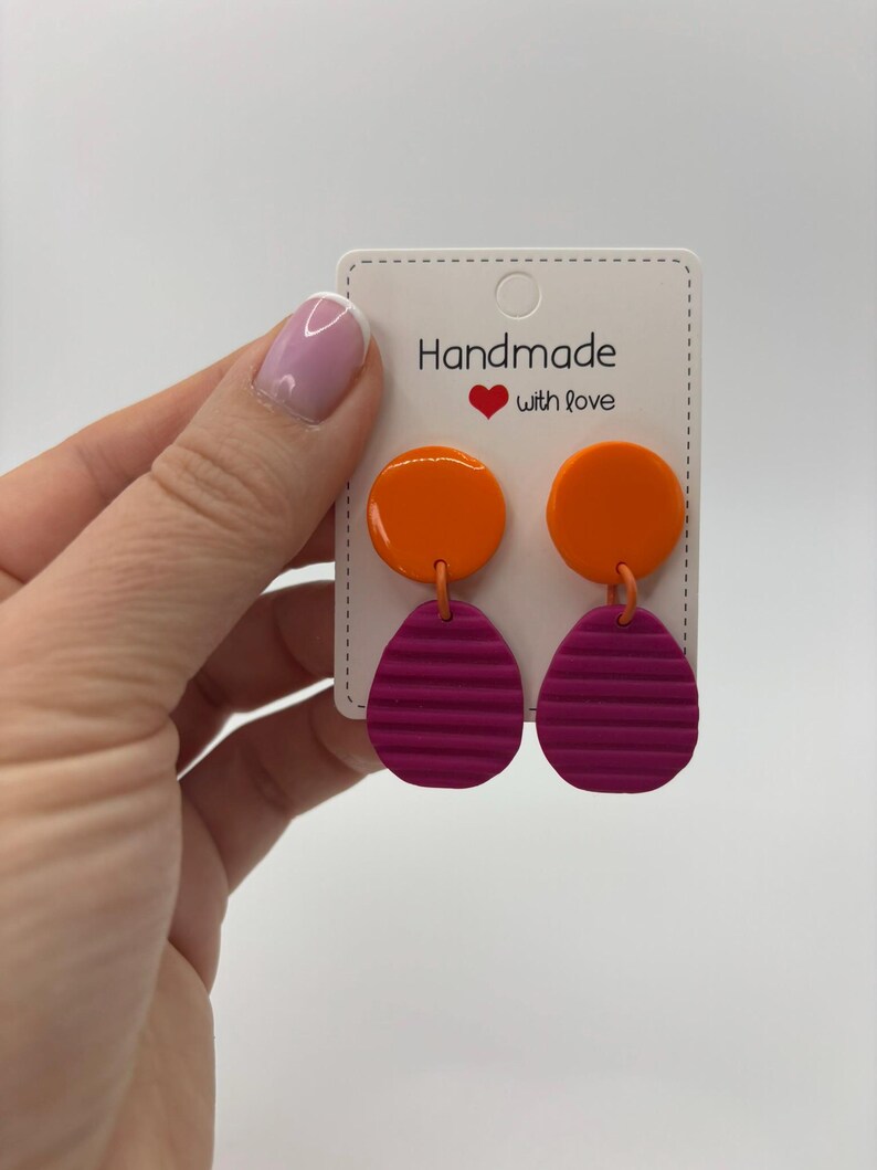 May include: Dangle earrings featuring a smooth, glossy orange circular top piece connected to a textured, matte magenta oval bottom piece with horizontal ridges. The earrings are attached to a white display card labeled Handmade with love.