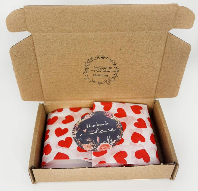 May include: An open brown cardboard shipping box containing white tissue paper printed with red hearts. A circular sticker with a floral design and the text "Handmade with love" is placed on top. The inside lid features a circular stamp with the text "HANDMADE with love polkabizze".