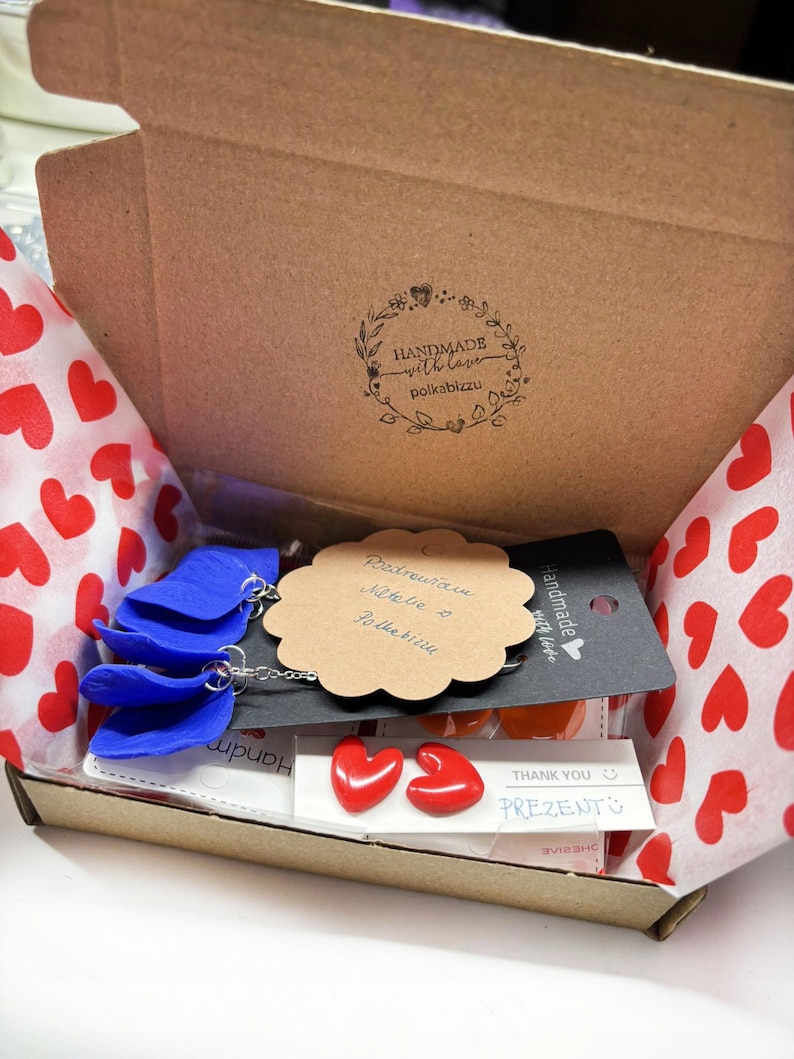 May include: A brown cardboard gift box lined with white tissue paper featuring red hearts. Inside are blue petal-shaped earrings on a black card, a scalloped tan gift tag with handwritten text, and red heart-shaped stud earrings on a white card.