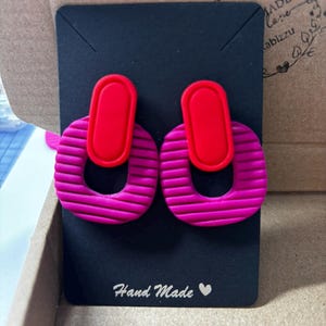Puede incluir: Un par de pendientes con una pieza superior ovalada de color rojo brillante conectada a un aro m&aacute;s grande de color magenta texturizado con estr&iacute;as horizontales. Los pendientes se presentan sobre una tarjeta negra con el texto Hand Made y un coraz&oacute;n.
