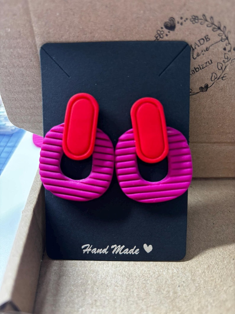 Puede incluir: Un par de pendientes con una pieza superior ovalada de color rojo brillante conectada a un aro m&aacute;s grande de color magenta texturizado con estr&iacute;as horizontales. Los pendientes se presentan sobre una tarjeta negra con el texto Hand Made y un coraz&oacute;n.