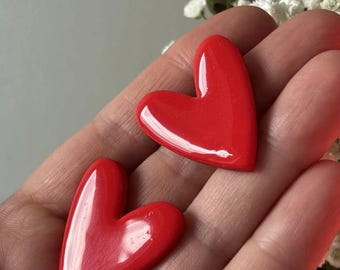 May include: Two small, glossy, bright red heart-shaped ornaments with a smooth, reflective surface. Each heart features a slightly asymmetrical, rounded design with a vibrant, solid red finish that catches the light, creating subtle highlights.