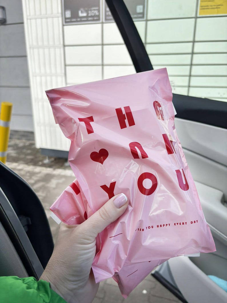 May include: A light pink plastic shipping mailer bag featuring the red text THANK YOU with a heart symbol replacing the letter A. The bottom right corner includes the smaller red text WISH YOU HAPPY EVERY DAY with two horizontal lines.