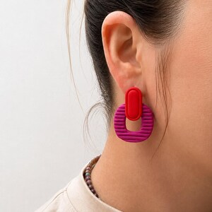Puede incluir: Un pendiente colgante que presenta una pieza ovalada roja lisa y s&oacute;lida conectada a un anillo circular magenta m&aacute;s grande y texturizado. El anillo magenta tiene ranuras horizontales en su superficie, creando un contraste con el &oacute;valo rojo.