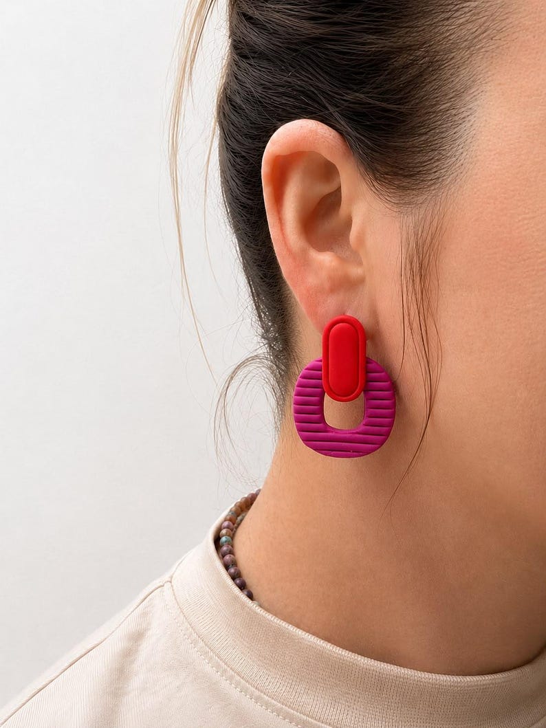 Puede incluir: Un pendiente colgante que presenta una pieza ovalada roja lisa y s&oacute;lida conectada a un anillo circular magenta m&aacute;s grande y texturizado. El anillo magenta tiene ranuras horizontales en su superficie, creando un contraste con el &oacute;valo rojo.