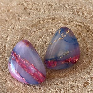 May include: A pair of triangular stud earrings featuring a smooth, polished finish with a marbled blend of lavender, blue, and deep pink hues, accented by embedded gold-coloured metallic flakes throughout the resin material.