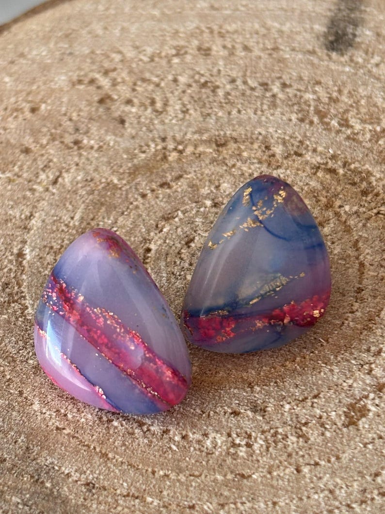 May include: A pair of triangular stud earrings featuring a smooth, polished finish with a marbled blend of lavender, blue, and deep pink hues, accented by embedded gold-coloured metallic flakes throughout the resin material.