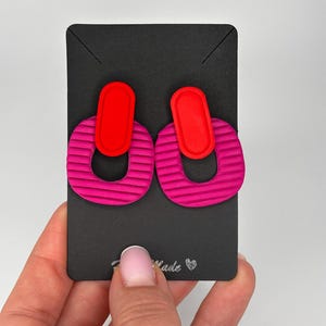 Puede incluir: Un par de pendientes que presentan una forma ovalada vertical de color rojo brillante unida a una pieza m&aacute;s grande en forma de U de color rosa magenta texturizado. Los pendientes se muestran en una tarjeta rectangular negra con el texto "Handmade" y un peque&ntilde;o icono de coraz&oacute;n impreso en la parte inferior.