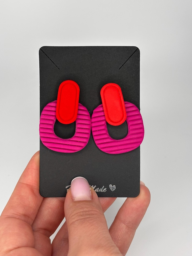 Puede incluir: Un par de pendientes que presentan una forma ovalada vertical de color rojo brillante unida a una pieza m&aacute;s grande en forma de U de color rosa magenta texturizado. Los pendientes se muestran en una tarjeta rectangular negra con el texto "Handmade" y un peque&ntilde;o icono de coraz&oacute;n impreso en la parte inferior.