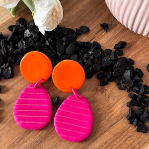 May include: A pair of dangle earrings featuring a bright orange circular top piece connected to a magenta teardrop-shaped bottom piece. The magenta sections have a horizontal striped texture across the surface of each earring.