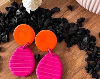 May include: A pair of dangle earrings featuring a bright orange circular top piece connected to a magenta teardrop-shaped bottom piece. The magenta sections have a horizontal striped texture across the surface of each earring.