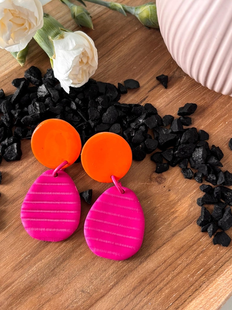 May include: A pair of dangle earrings featuring a bright orange circular top piece connected to a magenta teardrop-shaped bottom piece. The magenta sections have a horizontal striped texture across the surface of each earring.