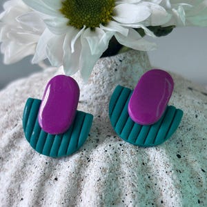 May include: A pair of handmade earrings. Each earring features a teal, semi-circular base with vertical lines, topped with a glossy, oval-shaped purple accent. The earrings are displayed on a textured, white surface, with a white flower in the background.