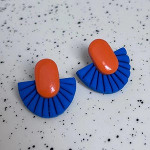 May include: A pair of stud earrings featuring a smooth, glossy orange oval top piece attached to a matte blue semi-circular base with a pleated, fan-like texture. The design combines a vibrant orange color with a bold blue, creating a distinct contrast in both shape and finish for these earrings.