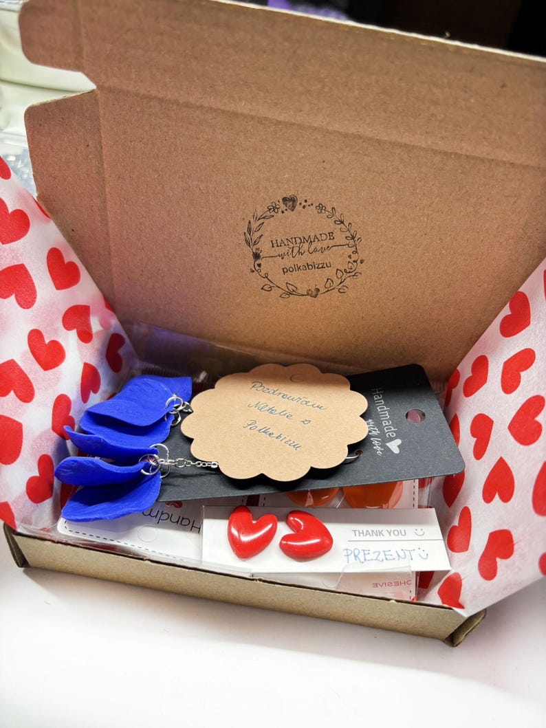 May include: A cardboard box lined with red heart-patterned tissue paper containing blue petal-shaped earrings on a black display card, a scalloped tan gift tag with handwritten text, and a pair of small red heart-shaped stud earrings on a white card.