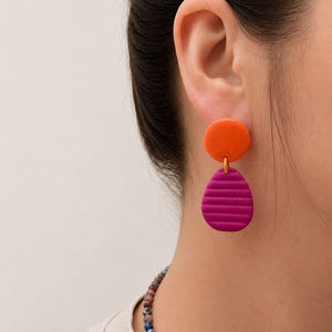May include: A drop earring featuring a smooth, circular orange stud connected to a larger, textured magenta pendant. The pendant has a teardrop shape with horizontal ridges across its surface. The earring is worn on an earlobe.