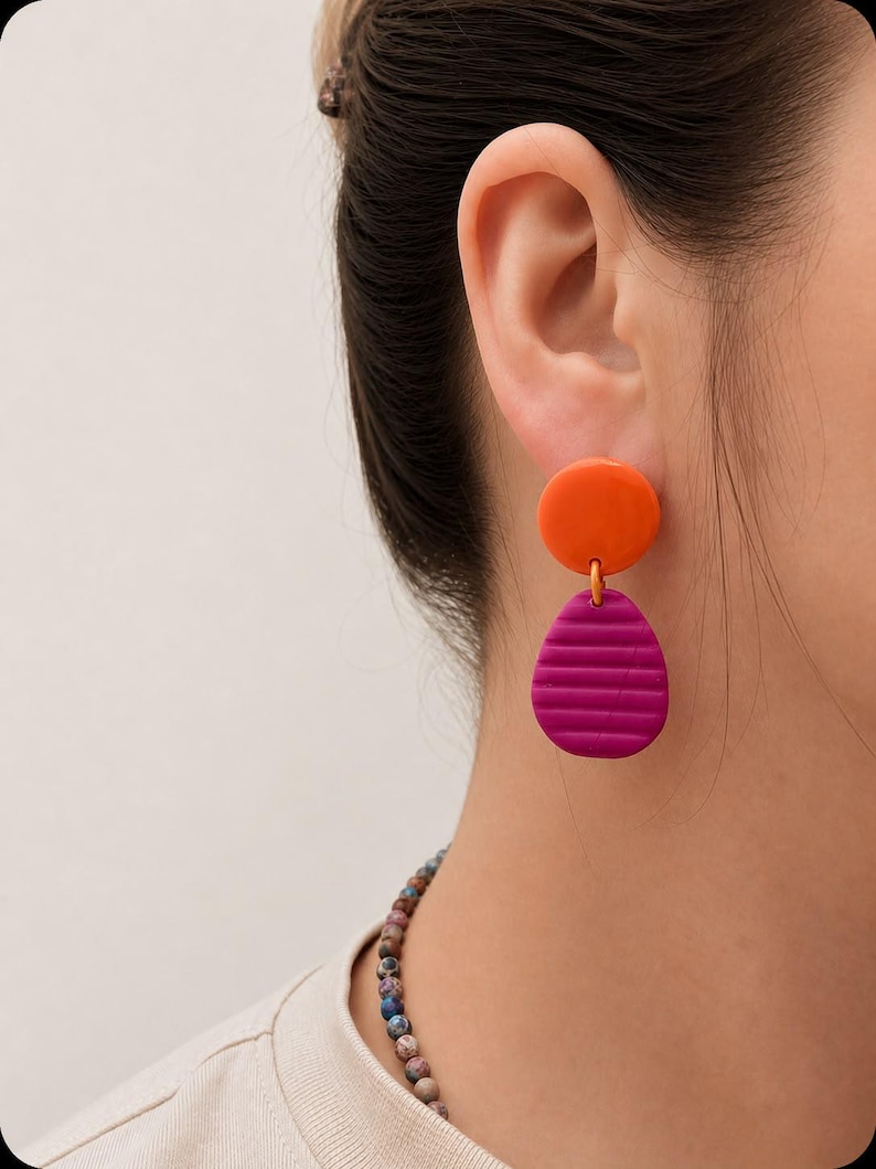 May include: A drop earring featuring a smooth, circular orange stud connected to a larger, textured magenta pendant. The pendant has a teardrop shape with horizontal ridges across its surface. The earring is worn on an earlobe.