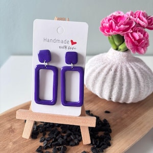 May include: A pair of handmade purple rectangular earrings displayed on a small wooden easel. The earrings are paired with a white card that reads "Handmade with love". A vase of pink flowers sits nearby.