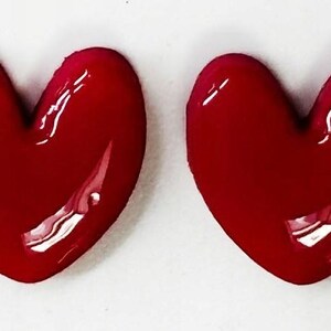 May include: Two identical heart-shaped objects with a smooth, glossy, deep red finish. Each heart features a rounded top and a pointed bottom, with a reflective surface that catches light, creating bright white highlights on the curved edges.