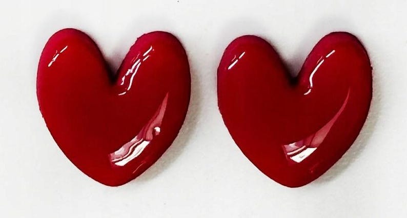 May include: Two identical heart-shaped objects with a smooth, glossy, deep red finish. Each heart features a rounded top and a pointed bottom, with a reflective surface that catches light, creating bright white highlights on the curved edges.