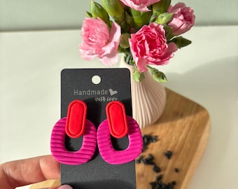 May include: A pair of dangle earrings on a black display card featuring the text Handmade with love. Each earring consists of a smooth red oval top piece connected to a larger, textured magenta oval bottom piece with horizontal ribbed lines.