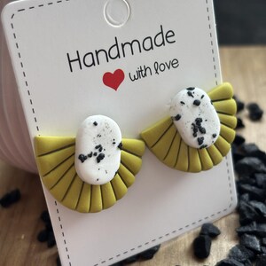 May include: A pair of handmade earrings with a fan-like design. The earrings feature a yellow base with a white oval center speckled with black dots. The earrings are displayed on a white card that reads "Handmade with love."