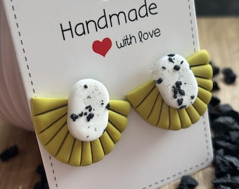 May include: A pair of handmade earrings with a fan-like design. The earrings feature a yellow base with a white oval center speckled with black dots. The earrings are displayed on a white card that reads "Handmade with love."