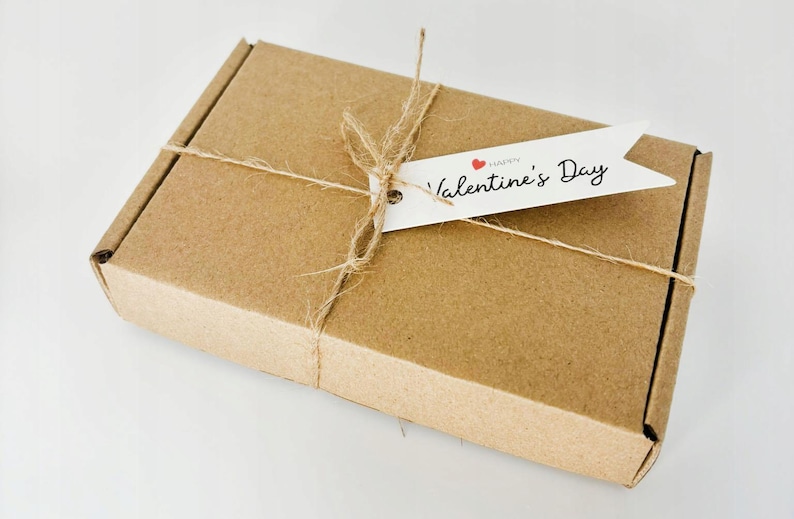 May include: A rectangular brown cardboard box tied with thin natural jute twine. A white paper tag is attached to the twine, featuring the black printed text Happy Valentine's Day and a small red heart icon above the word Valentine.