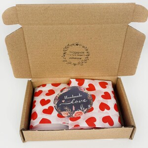 May include: A brown cardboard shipping box with an open lid featuring a floral wreath logo and the text "HANDMADE with love polkabizze". Inside, white tissue paper with red hearts covers the contents, topped with a round sticker reading "Handmade with love" surrounded by a floral and botanical illustration.