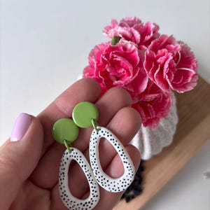 May include: A pair of lime green and white earrings with black polka dots. The earrings have a circular top and an oval-shaped bottom. The earrings are held in a hand, with pink flowers in the background.