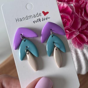 May include: A pair of handmade earrings with a three-layer design. The top layer is a curved, purple shape, followed by a teal leaf shape, and a beige teardrop shape at the bottom. The earrings are attached to a white card that says "Handmade with love."