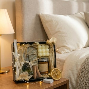 May include: A gift set in a black box, featuring a candle, diffuser, and other items. The candle is a light yellow color, and the diffuser has a clear bottle. The set includes cinnamon sticks and a dried orange slice. Fairy lights are wrapped around the box.