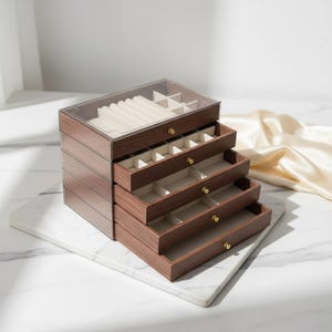 Może przedstawiać: Drewniane pudełko na biżuterię z wieloma szufladami, każda wyłożona miękką, kremową tkaniną. Pudełko ma przezroczysty wierzch i złote gałki. Widoczne usłojenie drewna. Pudełko stoi na białej marmurowej powierzchni. Wymiary w cm.