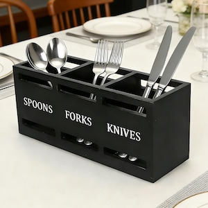 Puede incluir: Un soporte de utensilios de cocina de madera negra con tres compartimentos etiquetados como "SPOONS", "FORKS" y "KNIVES" en blanco. Contiene cucharas, tenedores y cuchillos. El soporte rectangular está sobre un mantel blanco.