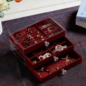 Könnte beinhalten: Ein klarer, dreistufiger Schmuck-Organizer mit rotem Samt-Innenraum. Die oberste Ebene hat Ringschlitze und Fächer, die mit Schmuck gefüllt sind. Die Schubladen darunter enthalten Halsketten, Armbänder und eine Uhr. Klare Kristallknöpfe zieren die Schubladen.