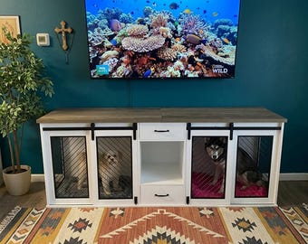 May include: A white and wood dog crate with sliding doors and black metal mesh. Two dogs are visible inside the crate. The crate is under a large television displaying an underwater scene. The room has a colorful rug.