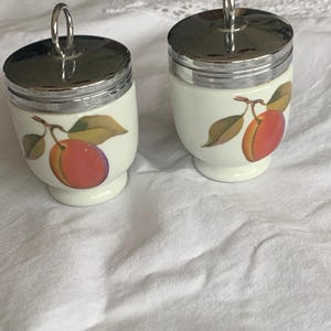 May include: Two white porcelain egg coddlers with a glossy finish, each featuring a colorful illustration of a single peach with green leaves on a branch. The containers have polished silver-tone metal lids with a circular ring handle on top.