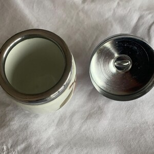 May include: A small white ceramic jar with a metallic silver rim and a matching circular metallic lid featuring a central handle. The jar has a smooth, cylindrical body and is shown from a top-down perspective, revealing its open interior.