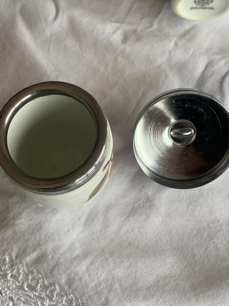 May include: A small white ceramic jar with a metallic silver rim and a matching circular metallic lid featuring a central handle. The jar has a smooth, cylindrical body and is shown from a top-down perspective, revealing its open interior.