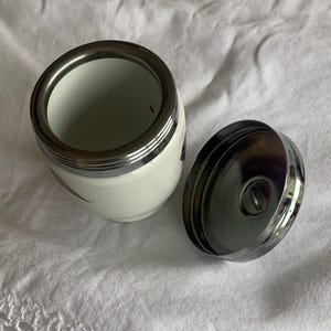May include: A small white ceramic jar with a metallic silver rim and a matching detachable silver lid. The jar features a smooth, rounded body and a wide opening, with a polished finish on the metal components and a clean white surface.