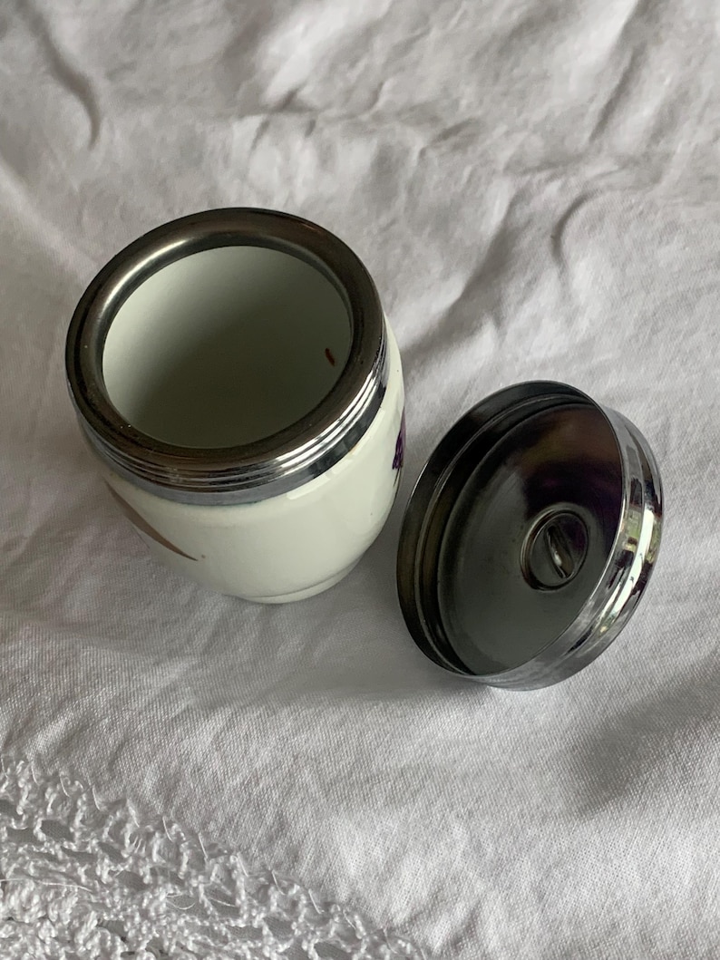 May include: A small white ceramic jar with a metallic silver rim and a matching detachable silver lid. The jar features a smooth, rounded body and a wide opening, with a polished finish on the metal components and a clean white surface.