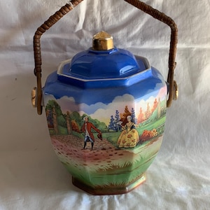May include: An octagonal ceramic jar with a blue lid and a gold-toned finial. The body features a colourful garden scene with two figures in period clothing. A brown woven handle is attached to the sides with gold-toned metal hardware.