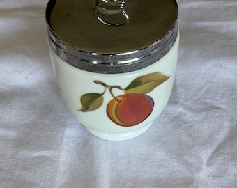 May include: A white ceramic egg coddler with a polished silver-tone metal lid featuring a circular ring handle. The body of the coddler is decorated with a colorful illustration of a single peach with green leaves attached to a small brown stem.