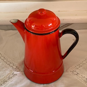 Op de afbeelding: Een feloranje geëmailleerde koffiepot met bijpassend deksel en een zwart handvat. De pot heeft een taps toelopend cilindrisch lichaam, een gebogen tuit en zwarte randdetails rond het deksel en bij de aanzet van het handvat.