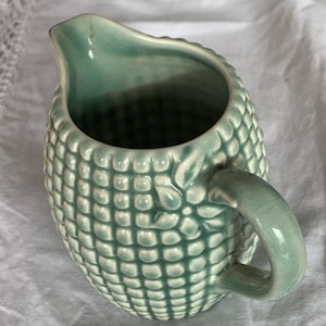 Può includere: Una lattiera in ceramica verde chiaro caratterizzata da una superficie testurizzata con un motivo a griglia ripetuto di quadrati in rilievo. La lattiera presenta un manico curvo, un beccuccio e una finitura smaltata lucida con crepe.