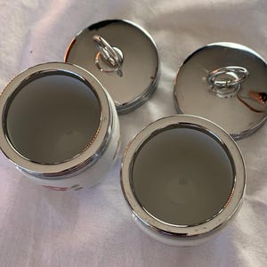 May include: Two white ceramic jars with silver-toned metal rims and matching circular lids. Each lid features a small, upright silver-toned ring handle. The jars have a smooth, white interior and a floral pattern visible on the exterior side.